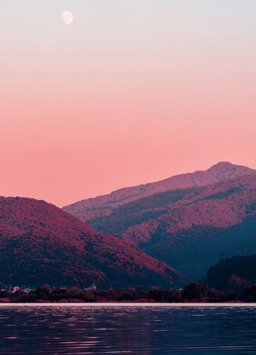 A wide shot of a lake with a pair of mountains in the backdrop, taken with a completely clear orange and blue sky, alongside a purple and red filter applied over the top