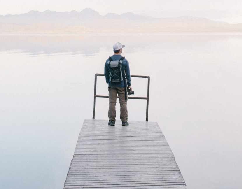 A young male photographer standing on a fishing deck, looking out onto a vast still lake with mountains barely visible in the foggy distance