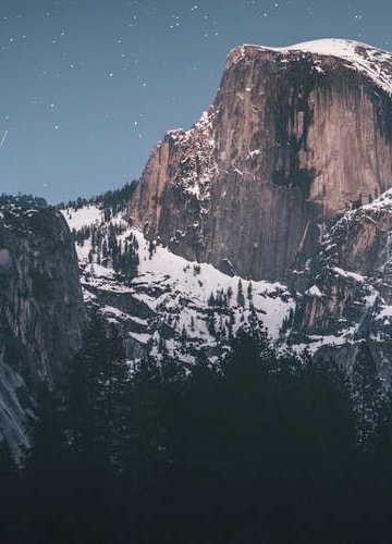 A wide shot of a single snowy mountain, housing an abundance of green trees scattered accross its surface, taken with a clear night sky where which multiple stars are visible