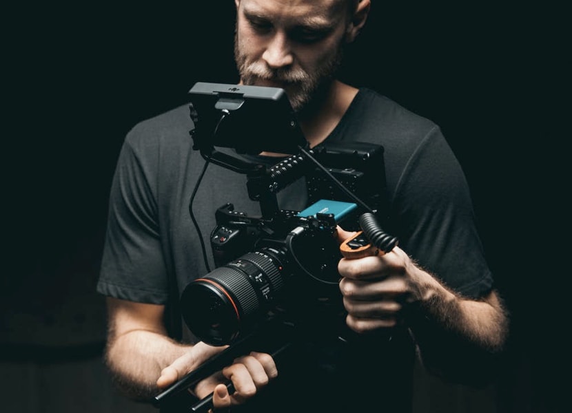 A male cinematographer wearing a dark-grey t-shirt, filming an object positioned off screen inside a dark room