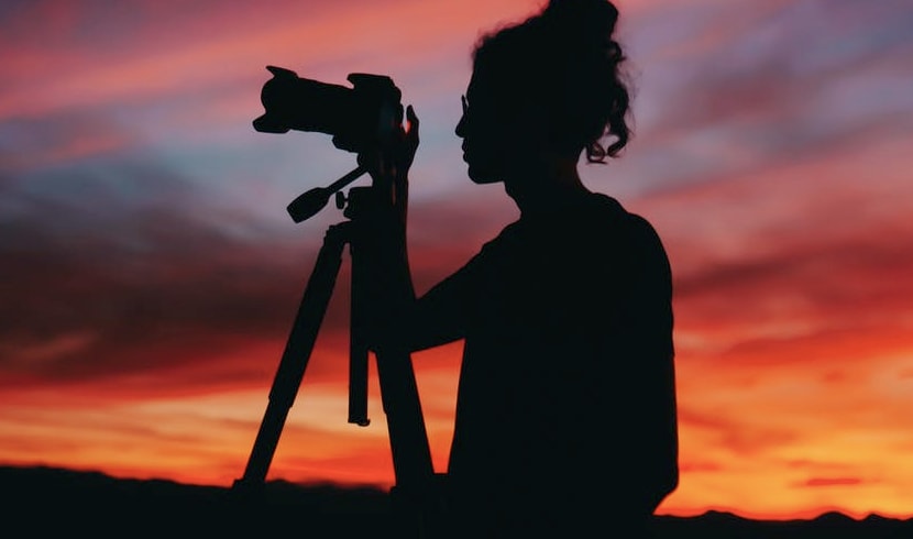 A middle aged female photographer looking through the lense of her DSLR camera positioned on a tripod, taken with a rich red and yellow sunset in the background