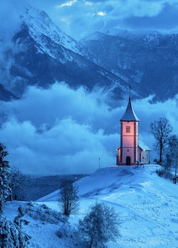 A wide shot of a snowy mountain range, taken at great heights with multiple clouds surrounding the mountains, alongside an illuminated church positioned alone and at the top