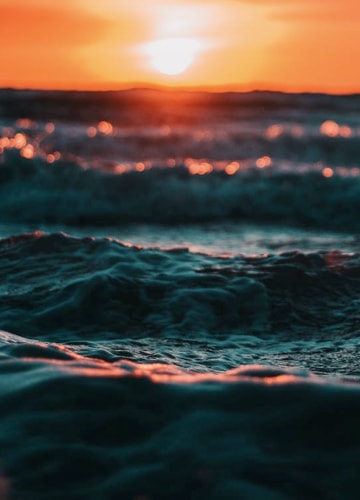 A close-up shot of the incoming waves rolling onto the beach shore, taken with a rich orange sunet in the background